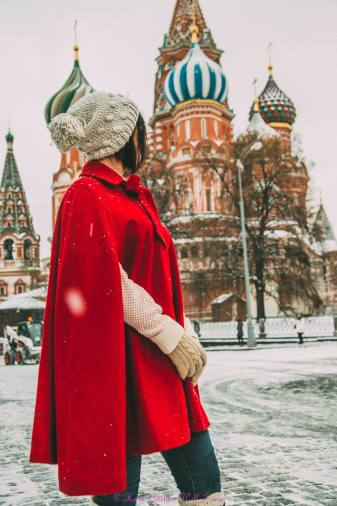 A woman poses on the wintery streets of Russia, posing with her back turned to the camera. She's wearing a red winter shawl and knitted hat.