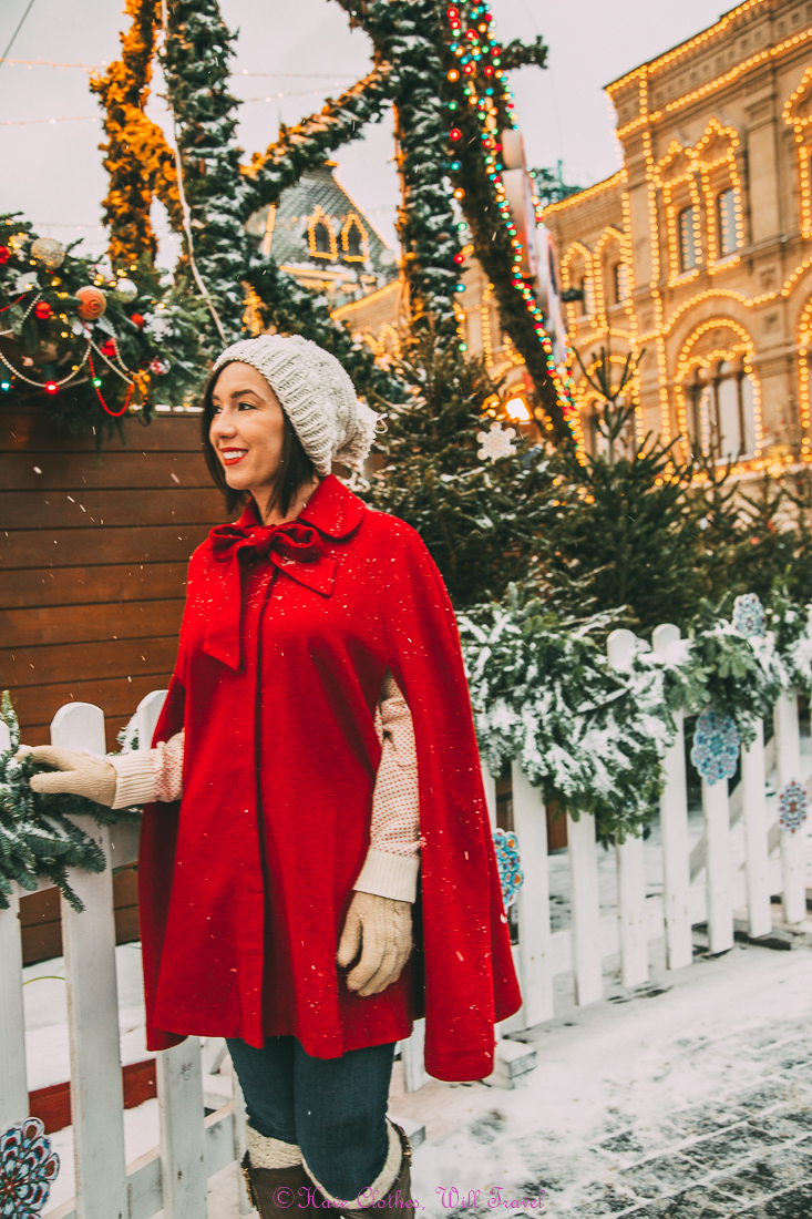 A woman is dressed in winter gear - a knit hat and red shawl overcoat, gloves, and jeans. She is standing next to a white fence decorated with pine wreathes and Christmas decorations, as the snow falls around her.