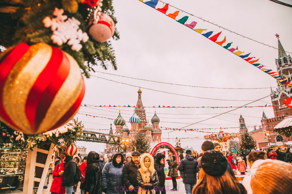 The bustling Moscow Christmas Market filled with visitors walking up and down the Christmas-decorated street. St. Basil's Cathedral's tall towers loom in the background.