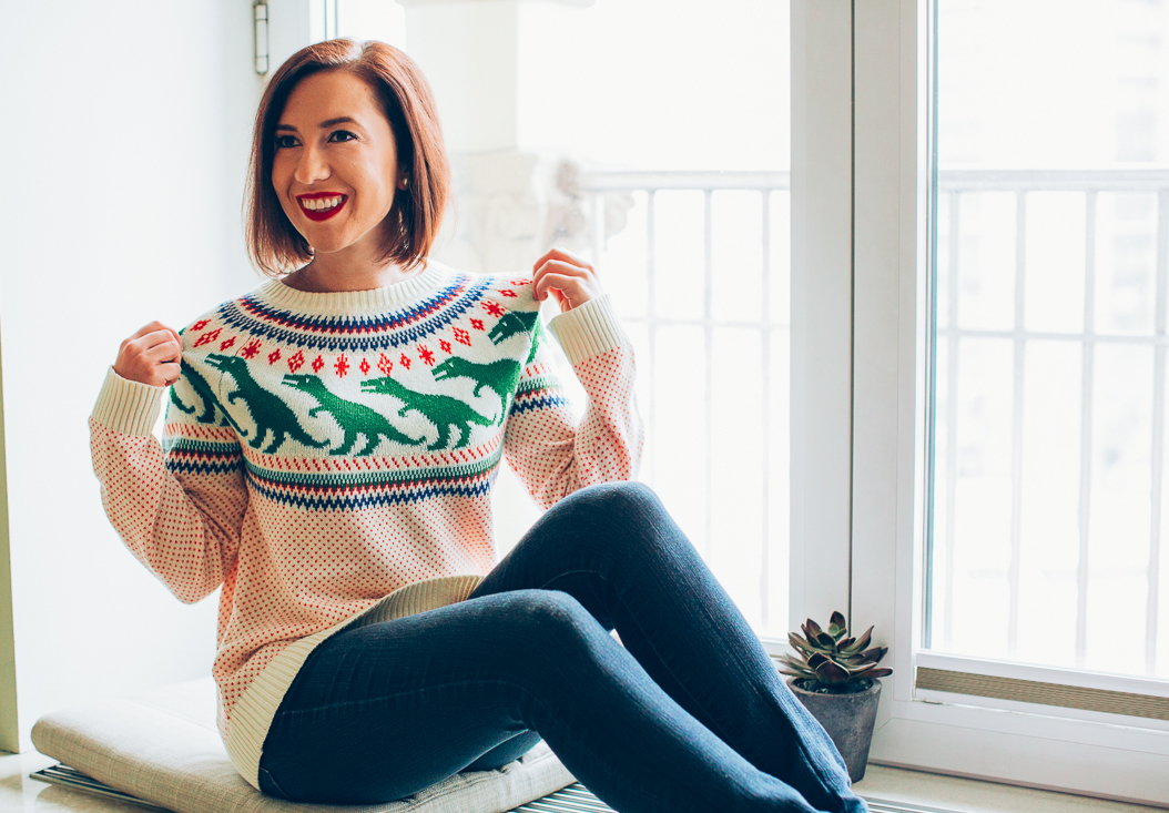 A woman poses in front of a bright window. She's wearing skinny jeans and a colorful sweater with a dinosaur pattern across the chest.