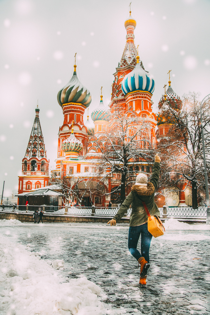 A woman wearing winter outer gear walks towards a colorful Russian building. Snow lines the cobblestone road and snowflakes fall around her.