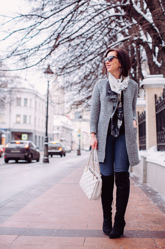 A woman poses while walking on a sidewalk. She's wearing knee-high boots, a long coat, scarf, and carrying a white handbag. There's snow on the sidewalk and tree branches behind her.