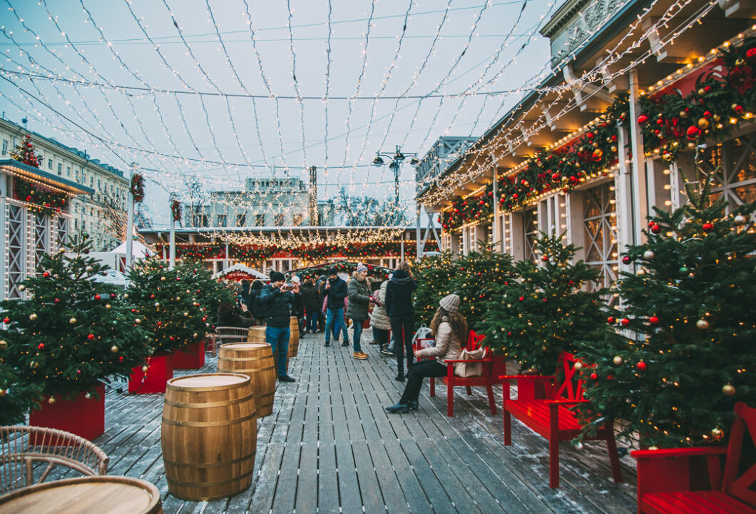 A Russian street market is filled with patrons. The market is decorated for Christmastime, lined with small Christmas trees, bright red benches, Christmas lights and garlands.