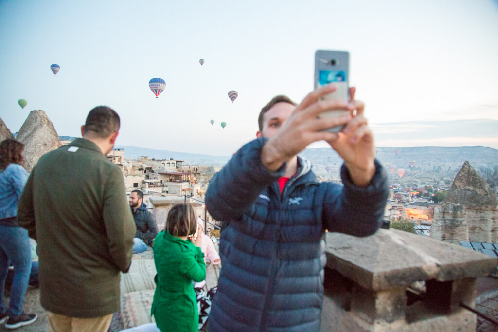 Tourists hold up phones and take selfies on a balcony in Cappadocia, Turkey, capturing the stunning views of hot air balloons rising in the sunset sky.