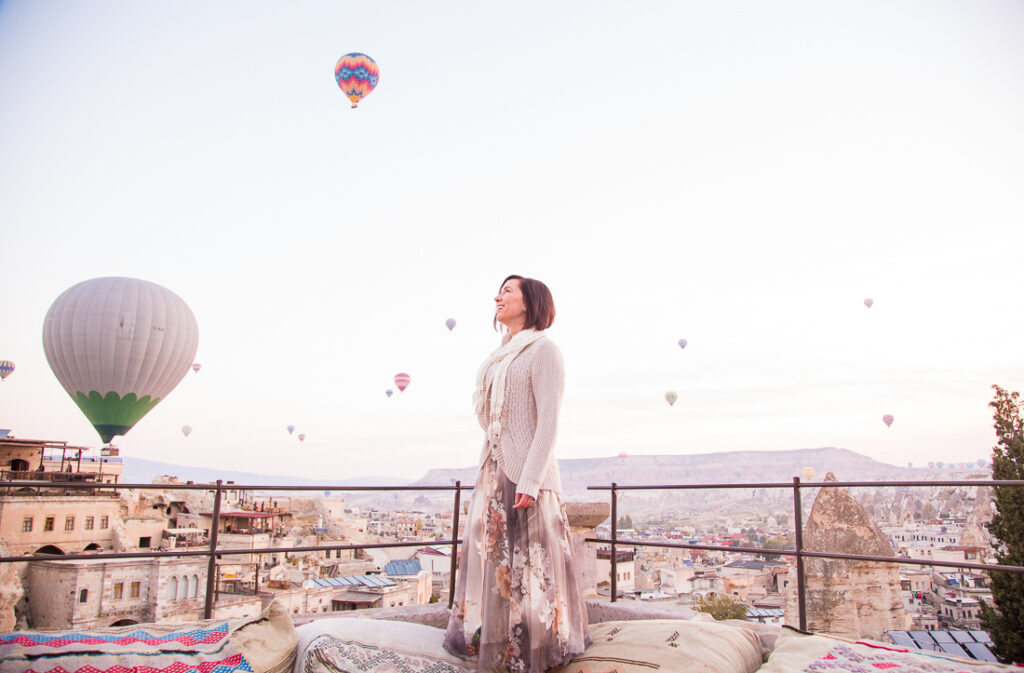 Lindsey of Have clothes, Will travel wearing a chiffon floral maxi skirt and tan cardigan, standing on a terrace with hot air balloons around her in Cappadocia, Turkey