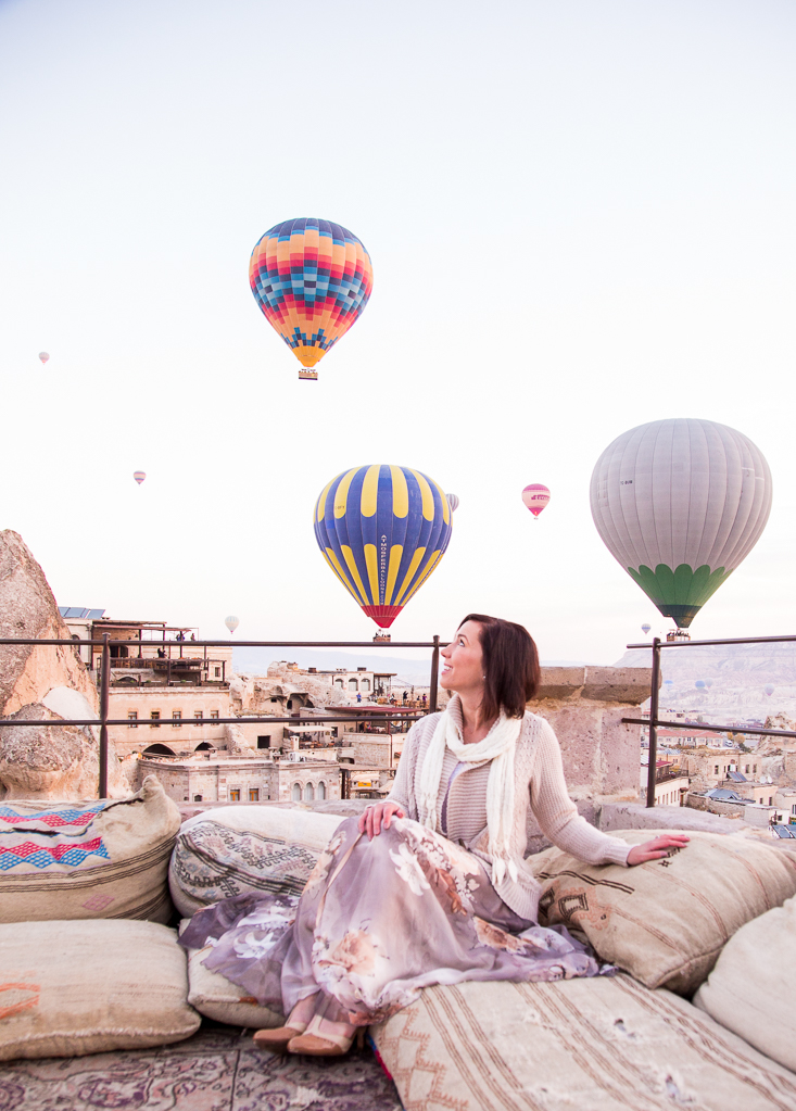 A woman sits on a balcony in Cappadocia, Turkey as hot air balloons rise in the sky behind her.
