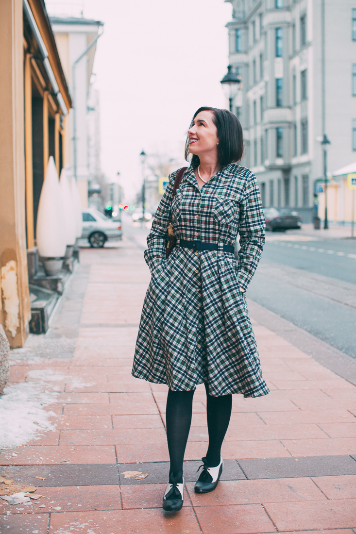 A woman walks down a sidewalk, window shopping. She's wearing a knee-length black and white flannel dress with black tights.