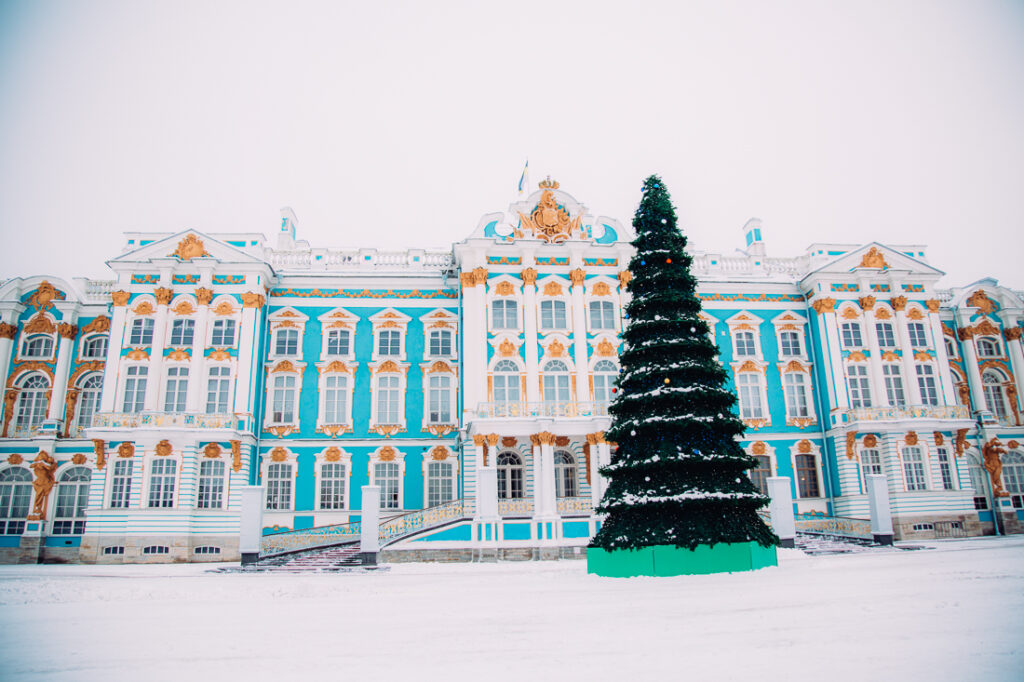The exterior of Catherine Palace on a wintery day in Russia. The grounds are covered with snow, there's a giant pine tree in front of the building.