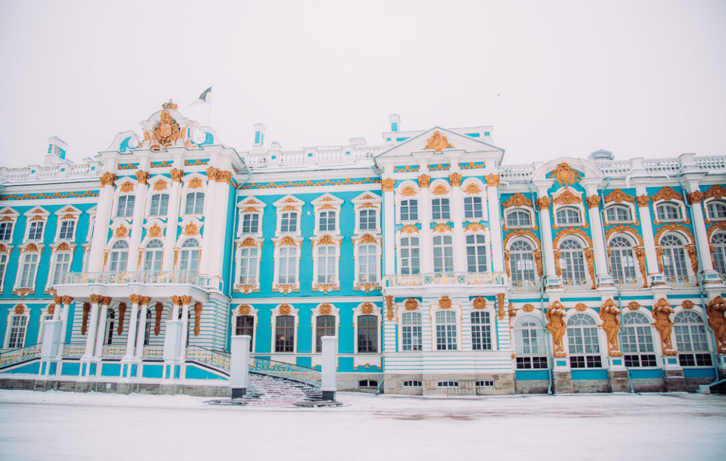 The outer front façade of Catherine Palace in Pushkin, Russia is turquoise and white with gold details, and the grounds in front of the Palace are covered in white snow.