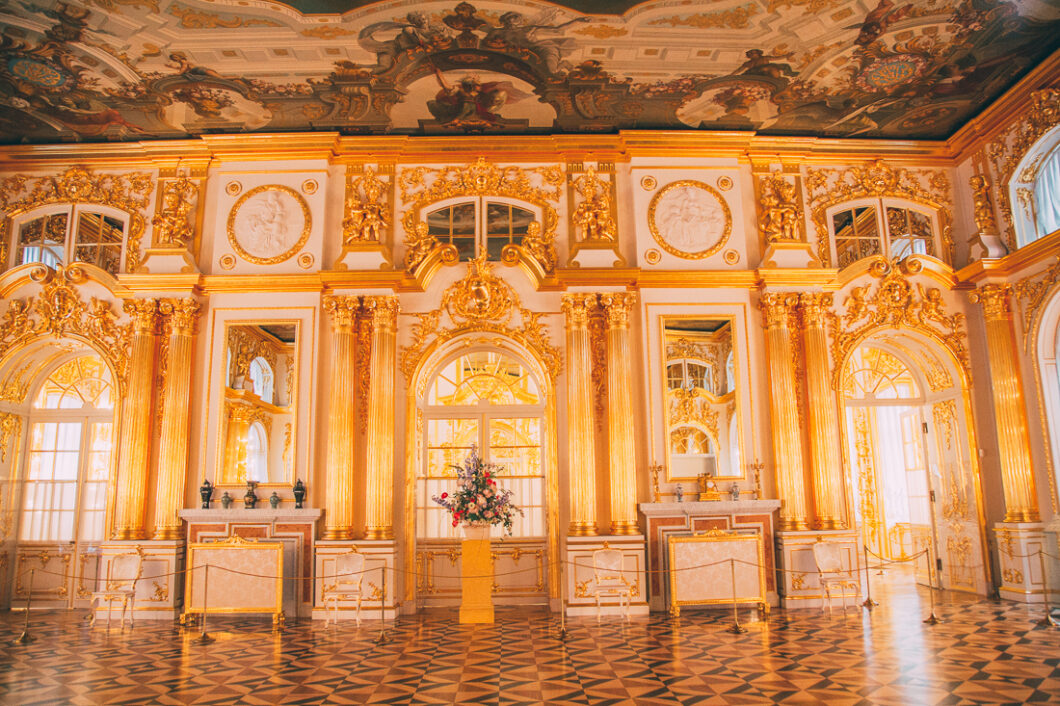 A stunning gilded gold room inside Catherine Palace in St. Petersburg, Russia. The room has high ceilings adored with crown molding, high archway windows, parquet wood floors, and stunning décor.