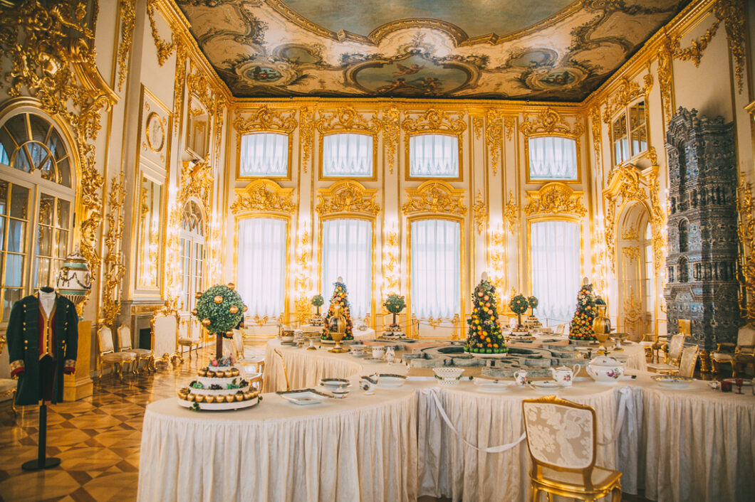 An interior image of a large dining room inside Catherine Palace. The grand room features white walls and windows adorned in gilded gold molding, crown-molding covered ceilings, and elegant dining tables set with ornate china.