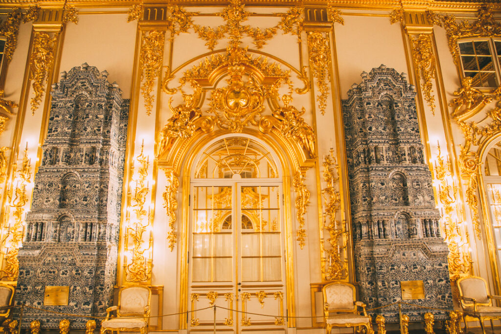 A single wall inside Catherine Palace, adorned with gilded gold molding surrounding an arched doorway, and two giant fireplaces on either side of the doorway.