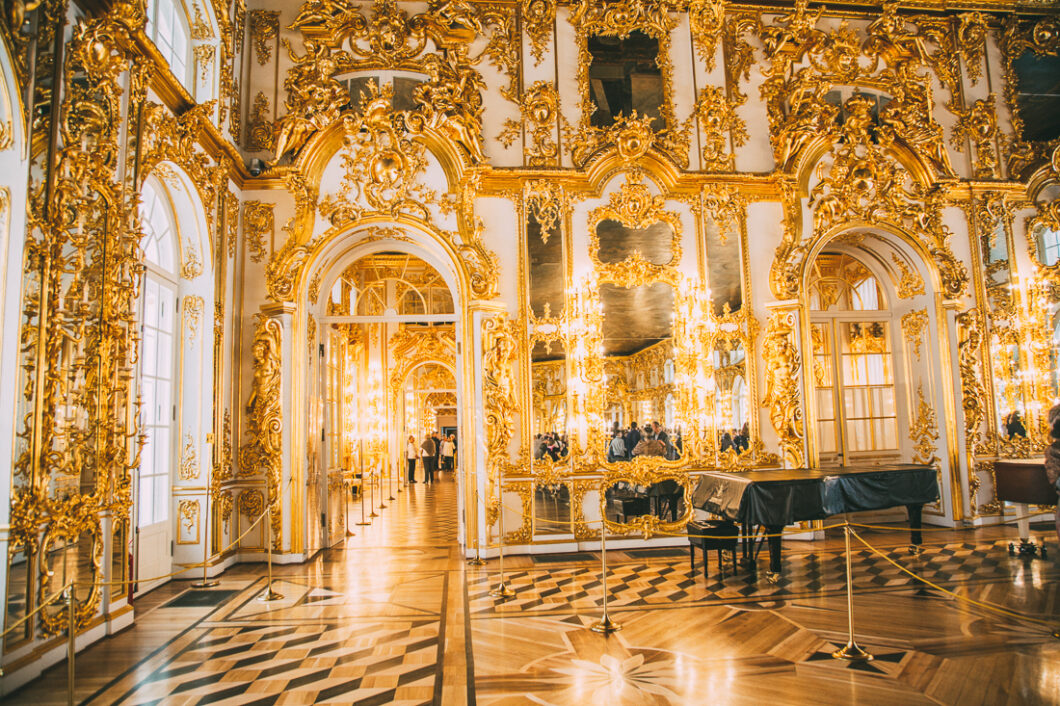 An interior image of another grand gilded-gold room inside Catherine Palace. Ornate gold molding covers the walls and surrounds mirrors, archways, and windows. Crowds of tourists gather to see tour the palace.