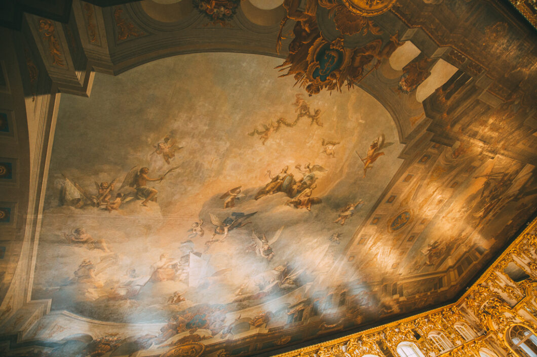 A giant hand-painted mural expands across a tall ceiling inside Catherine Palace in Russia. The mural depicts a heavenly scene with angels and nymphs dancing among the clouds.