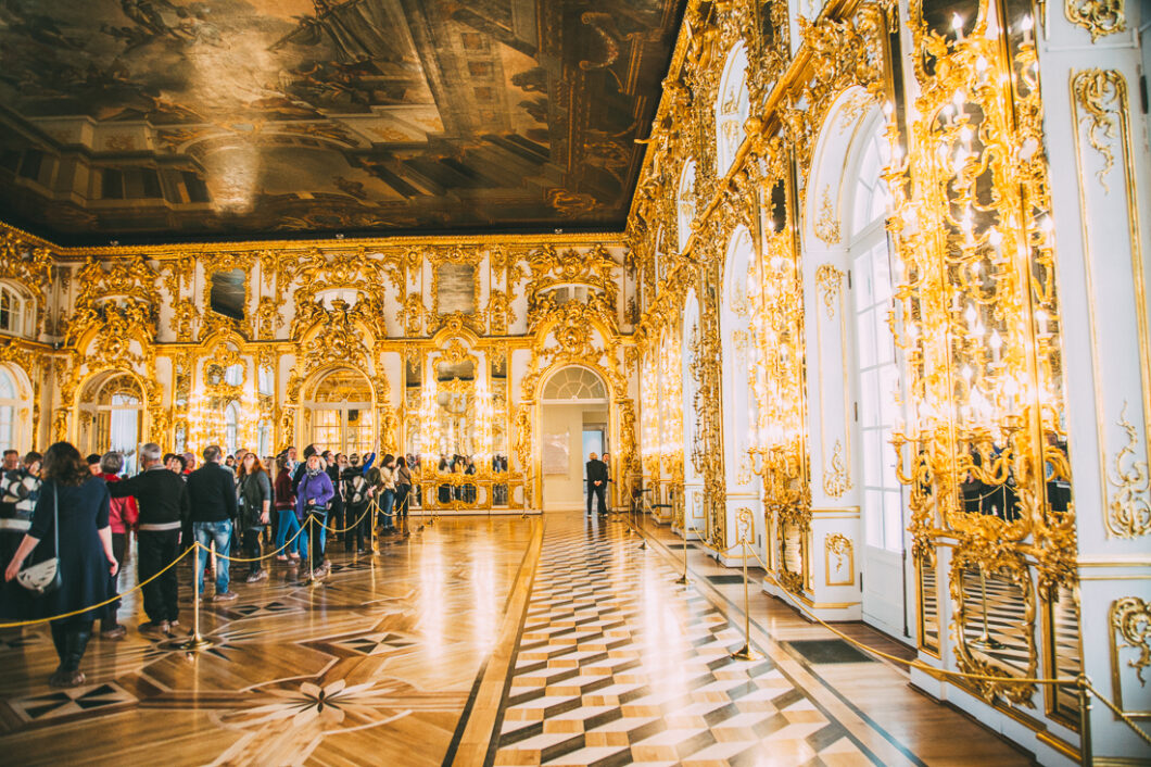 Tourists line up inside a grand room in Catherine Palace, waiting for a tour. The large room has parquet wood floors, gilded gold walls, and high ceilings covered in elegant crown molding.