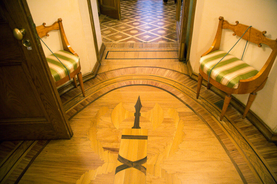 Stunning patterned inlay wooden floors in a hallways inside Catherine Palace. There are two decorative chairs against either wall in the small oval-shaped hallway.