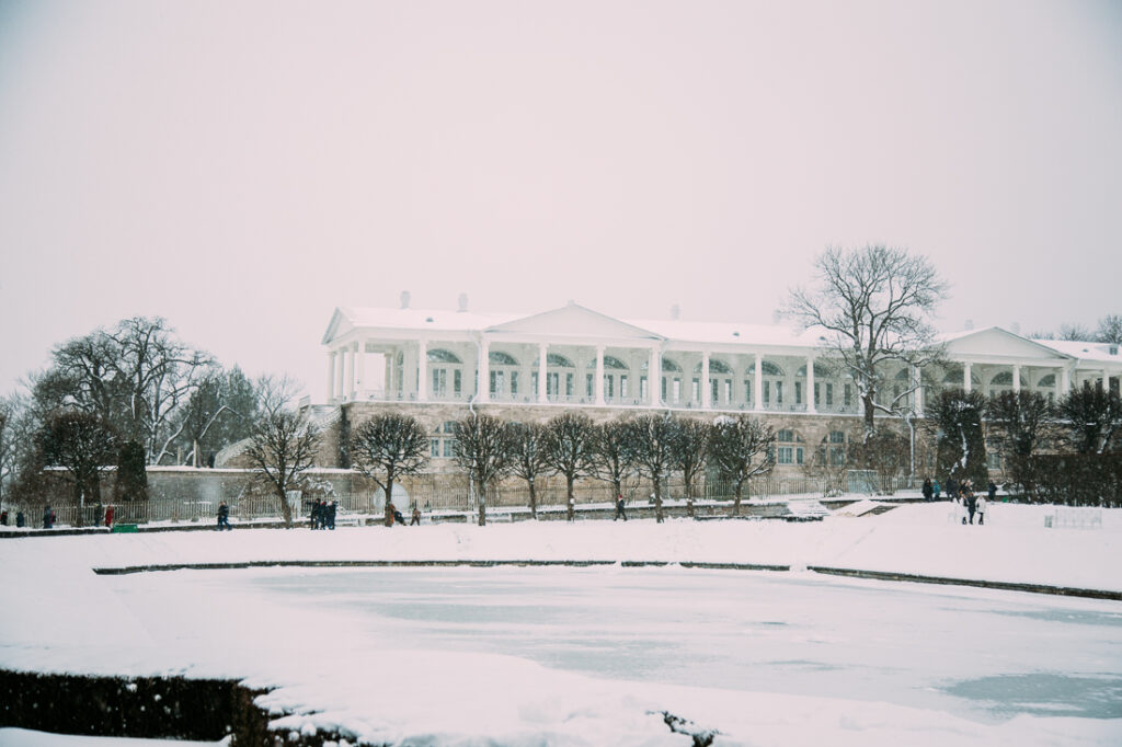 An outdoor image of a snowy day in Russia, with snow-covered grounds and an iced-over pond and snow-covered building in the background.
