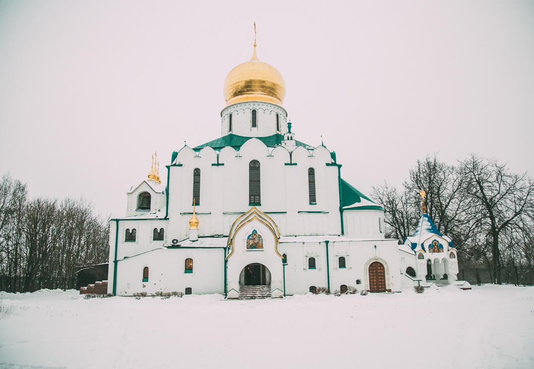 The exterior Feodorovskiy Gosudarev Cathedral in Russia on a snowy winter day. The large building is mostly white with green roof detailing and a giant gold obelisk at the very top. The grounds in front of the cathedral are covered in snow.