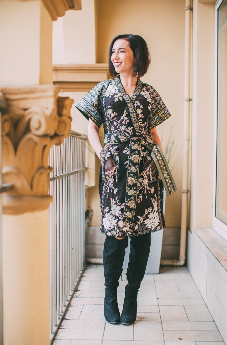 A woman stands on a balcony, modeling a knee-length wrap style kimono-inspired dress. The dress is black and has all-over floral embroidery with a tied waistband. She is also wearing knee-high black boots.