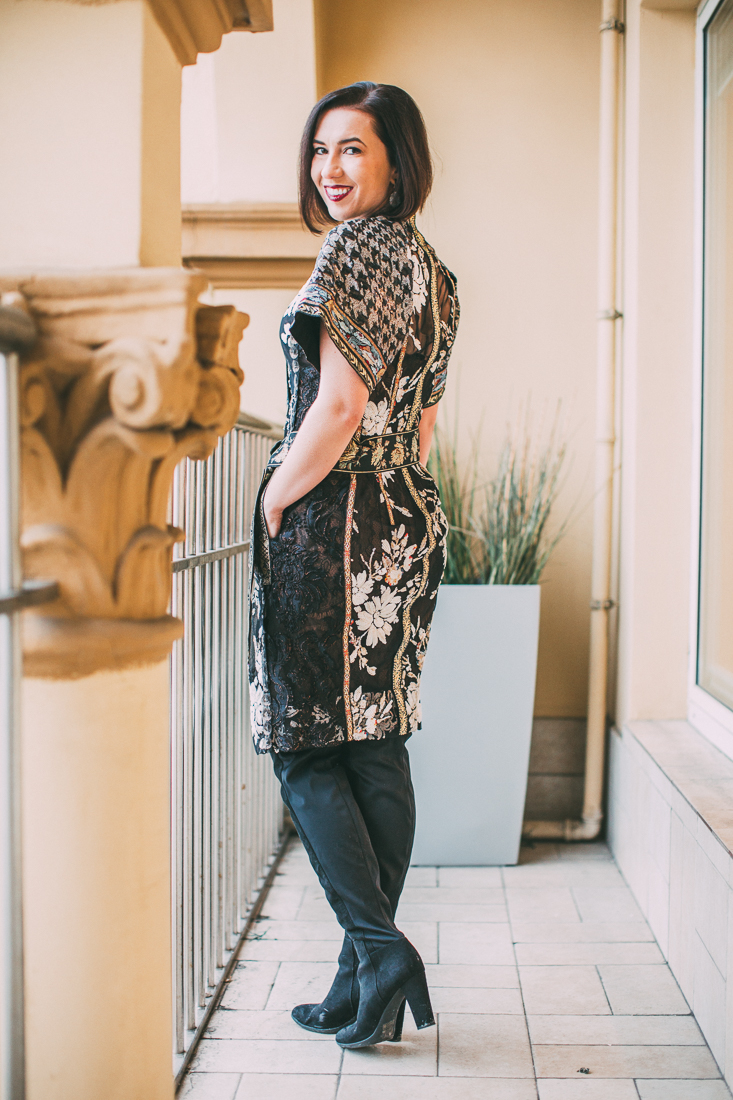 A woman poses turned away from the camera, looking over her shoulder. She wears a knee-length black wrap dress that's covered with gorgeous kimono-style floral patterns.