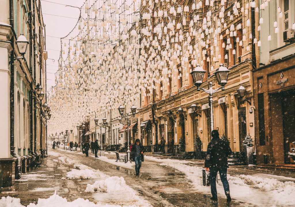 An image shows a Russian street in winter, with snow on the ground, pedestrians walking the streets, and a canopy of hanging lanterns over the road.