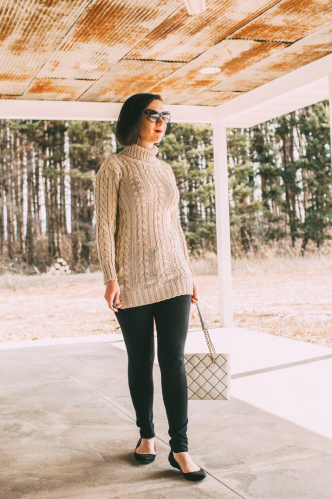 Front Pose of a women wearing sweater dress, black pants, holding a white bag and black pumps shoes