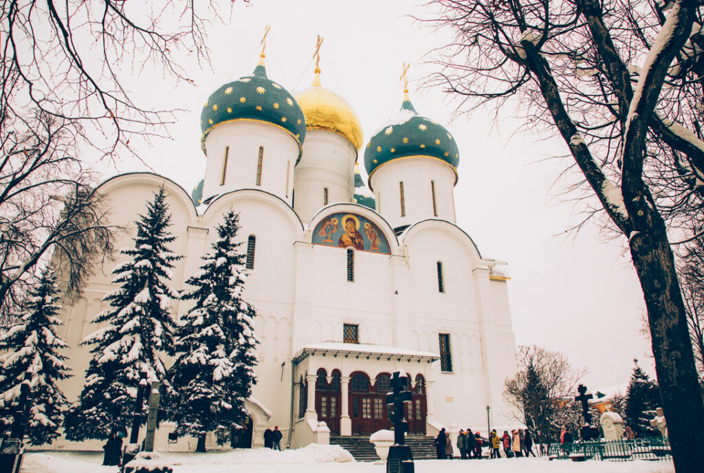 Sergiyev Posad, an Orthodox cathedral in Russia. Tourists mull around in front of the large white building. There's snow on the ground and snow covered trees.