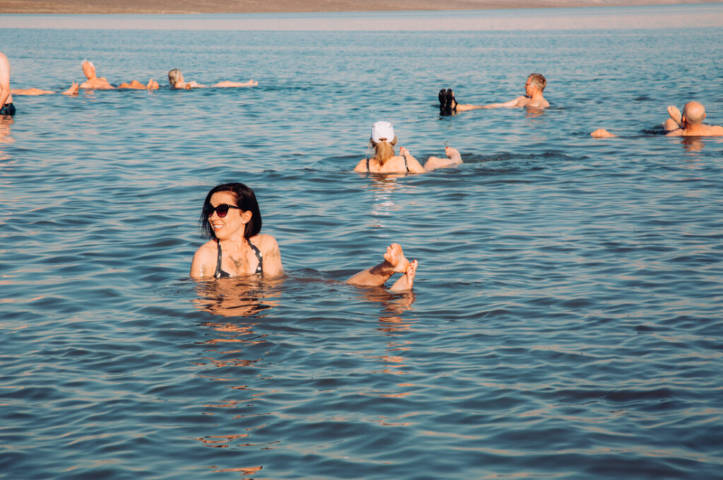 A young women with short brown hair and sunglasses floats in a body of water, surrounded by other tourists. Her head, shoulders, and feet are floating above the Dead Sea water, and she's looking over her shoulder, smiling.