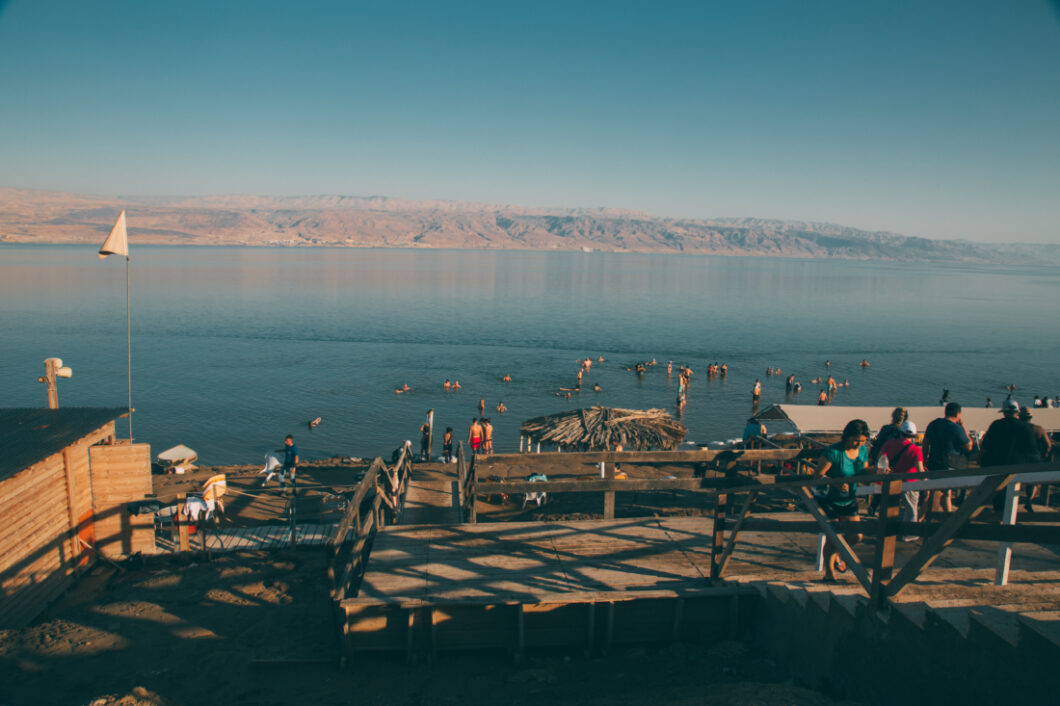 A photo of the shoreline and beach of The Dead Sea. Tourists wade in the water and fill the beach. Rocky landscape rises in the background against a hazy blue sky.