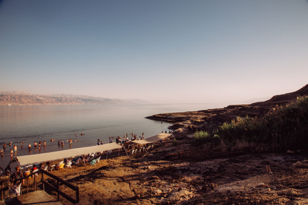 A stunning landscape photograph of the Dead Sea and surrounding mountains. Tourists line the shoreline and wade into the waters of the Dead Sea. The sky above is light blue and slightly hazy.