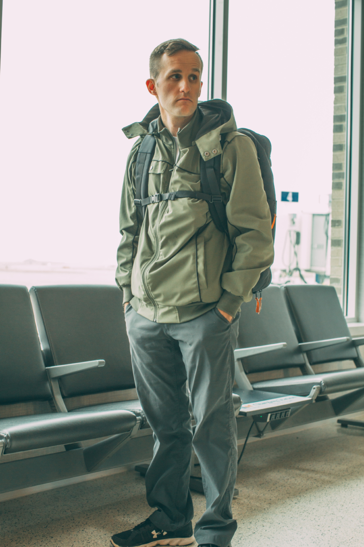 Zac wearing a green jacket with grey travel pants and a backpack standing in an airport
