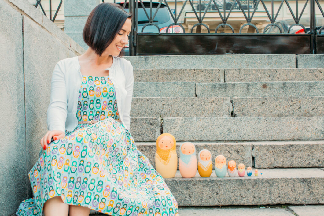 A woman sits on stone steps, posing next to a set of Russian nesting dolls set up on the stair beside her. She's wearing a blue dress with a cartoon nesting doll pattern and a white cardigan.