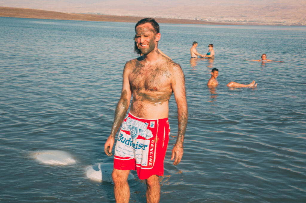 A man stands at the shoreline of the Dead Sea, with the expansive body of water behind him. He's wearing red and white Budweiser Beef shorts, and has mud smeared on his arms, upper body, and face. His eyes are squinted in the sunlight.