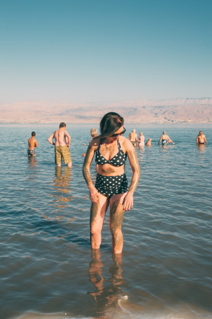 A woman with short brown hair, sunglasses, and a black and white polka dot bikini stands in shallow water on the shore of the Dead Sea. Her arms, legs, and upper body is covered in Dead Sea mud.