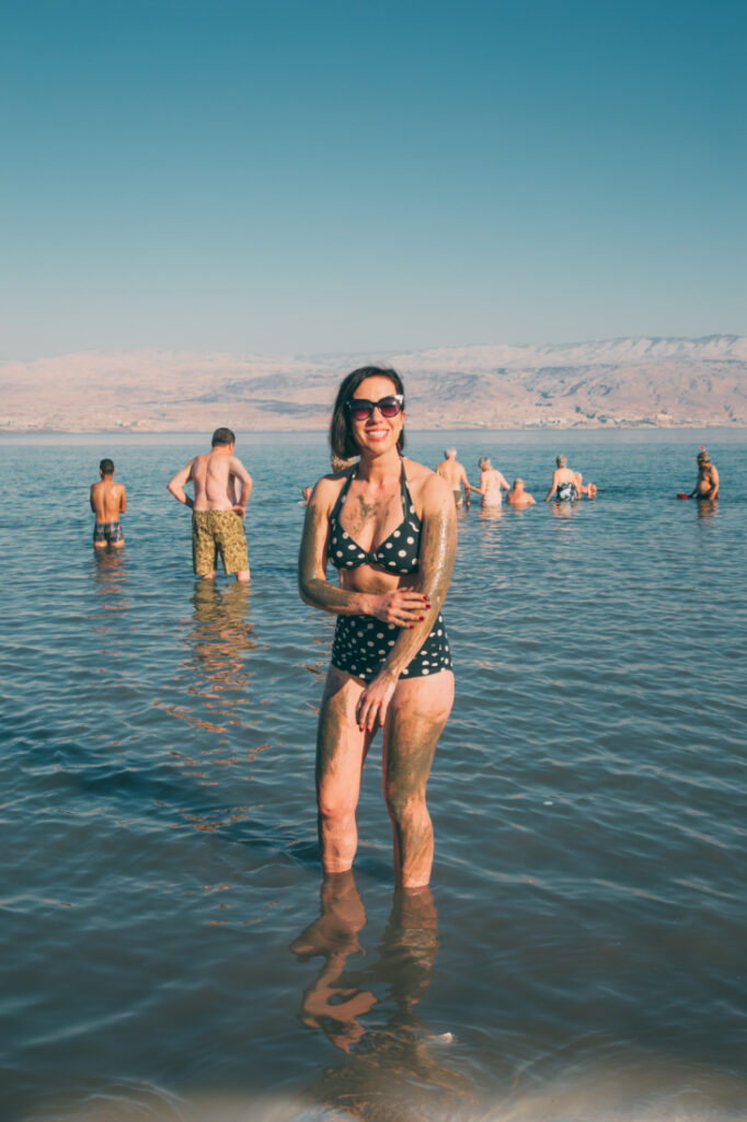 A woman wearing a black and white polka dot bikini stands in almost knee-high water on the shore of the Dead Sea. She is smearing Dead Sea mud on her arms, and is smiling at the camera.