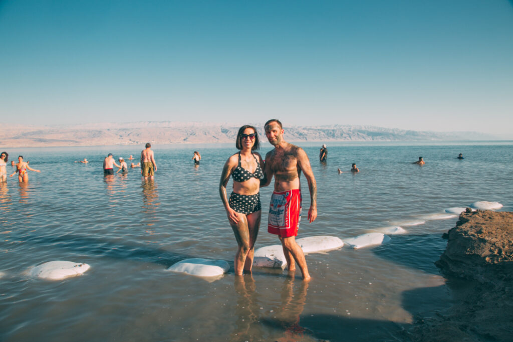A man and woman pose together on the shoreline of the Dead See - an expansive body of water with mountains in the background. They're standing in ankle-height water. Other tours stand in the water behind the couple.