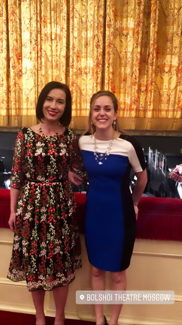 Two women pose together in front of the Historic Stage at the Bolshoi Theater. The woman on the left is wearing a white, red, and black floral patterned dress. The woman on the right is wearing a blue, white, and black color block dress.