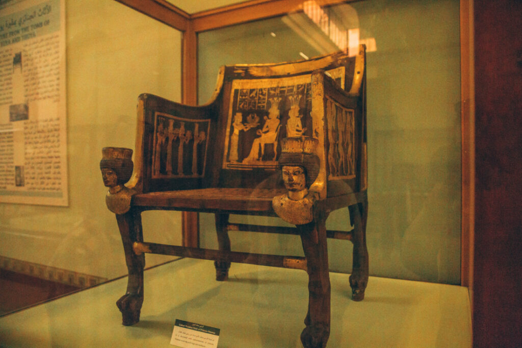 A large wooden chair, adorned with gold inlay and ornate carvings, sits inside a glass display case at the Egyptian Museum.