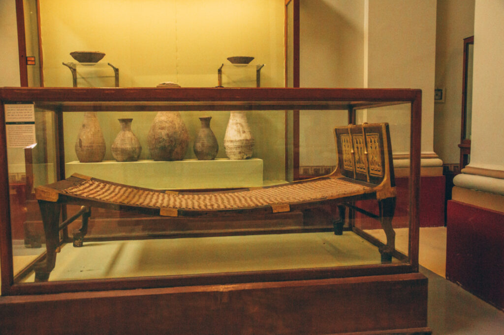 An ancient bed - a small wooden frame with a lang piece of cloth stretched over the frame, stands inside a glass display case at the Egyptian Museum.
