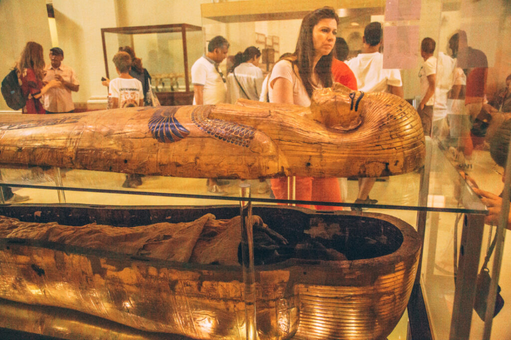 A mummy lays inside an opened sarcophagus displayed in a clear glass display case. The lid of the sarcophagus rests over the base on a raised plane of glass. 