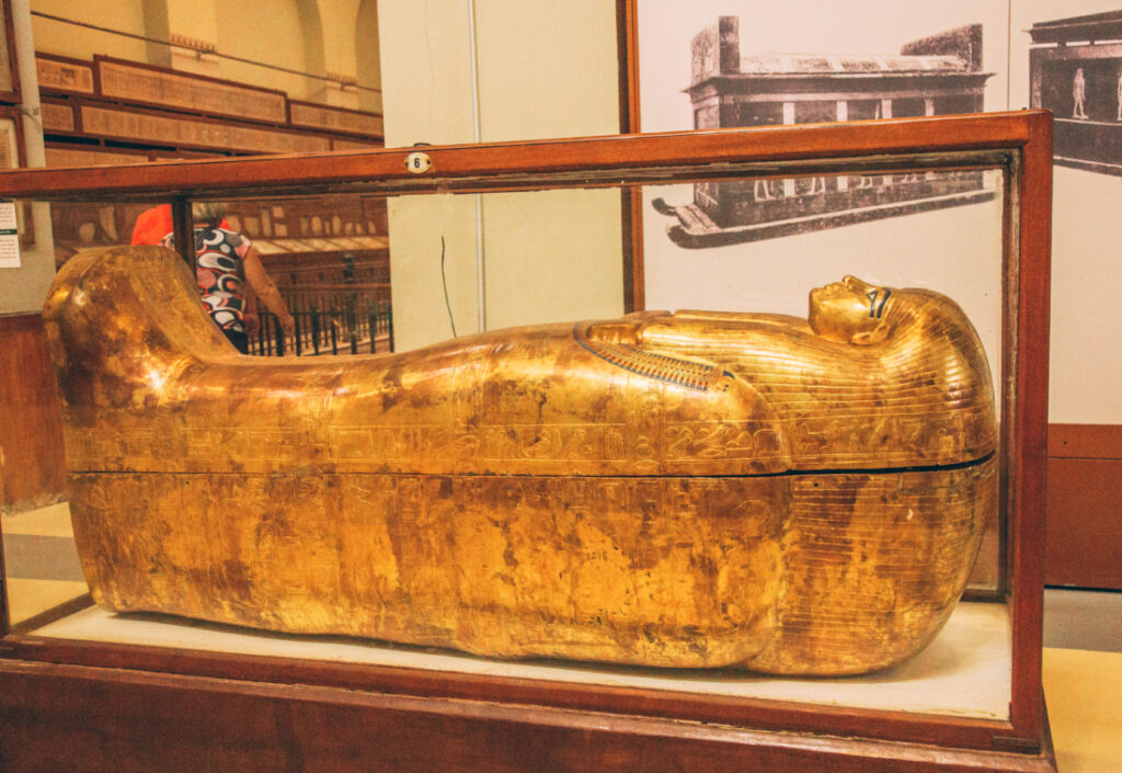 A large all-gold sarcophagus with ornate carvings sits inside a wood-framed glass display case at the Egyptian Museum.