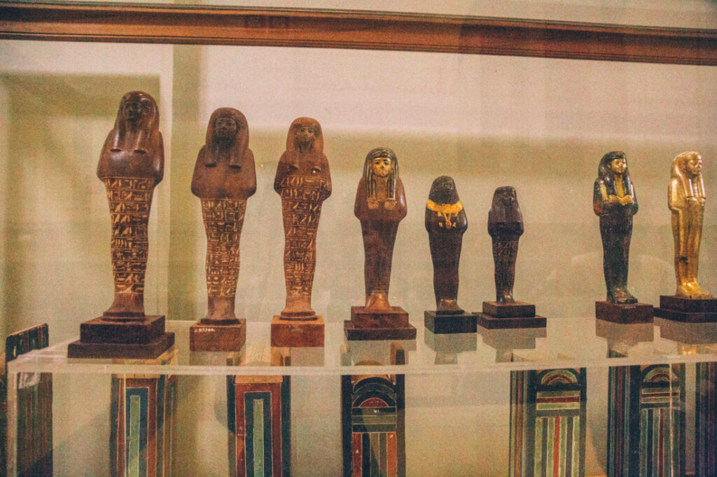 Small Egyptian statues carved from wood and stone stand in ascending order of height from left to right on glass shelf inside a display case at the Egyptian Museum.