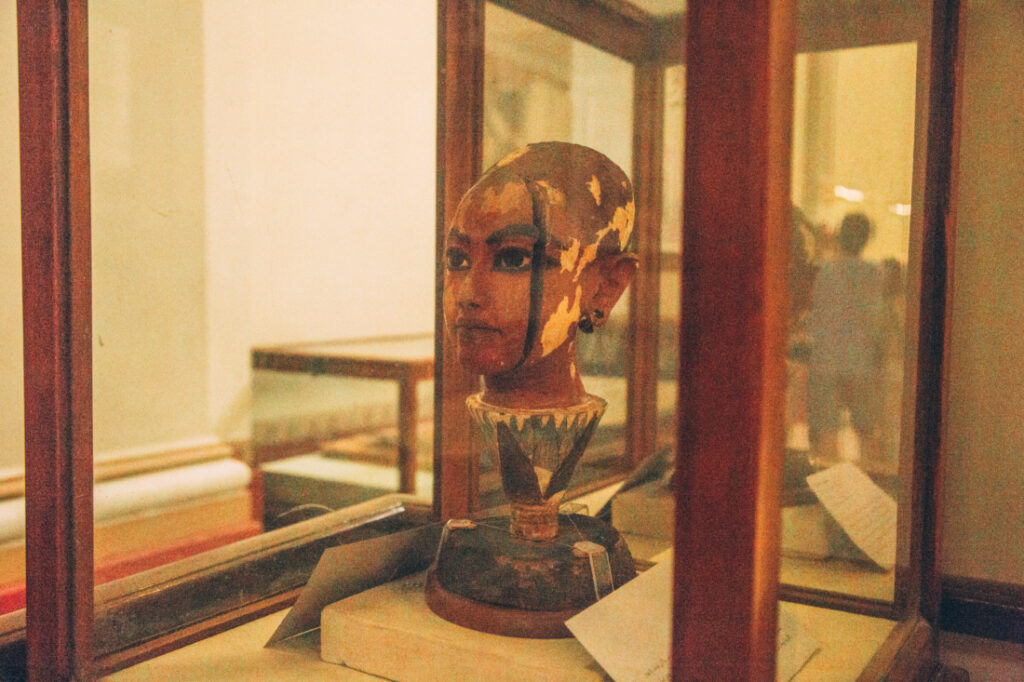 A statue of young King Tut's face carved from wood stands on a display stand inside of a glass case.