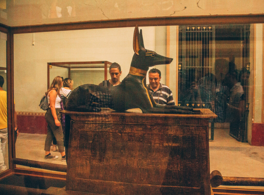 A statue of an Egyptian dog carved from black stone and painted with gold accents sits atop an ancient wooden box inside of a glass display case at the Egyptian Museum.