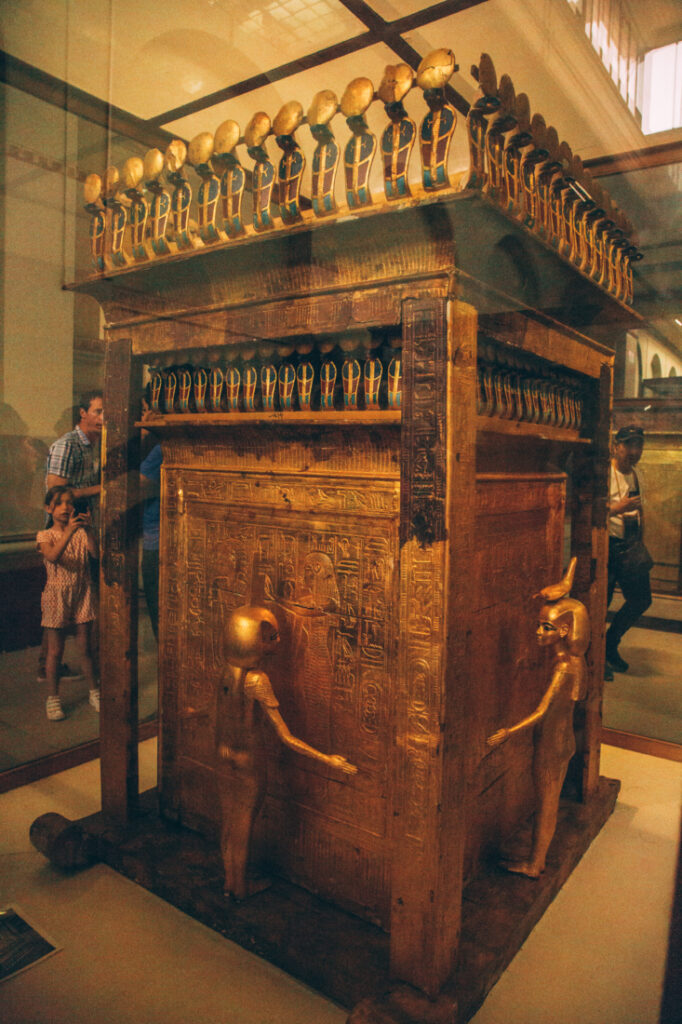 A giant sculpture carved from wood and stone, adorned in ornate carvings and statues, sits inside of a display case at the Egyptian Museum.