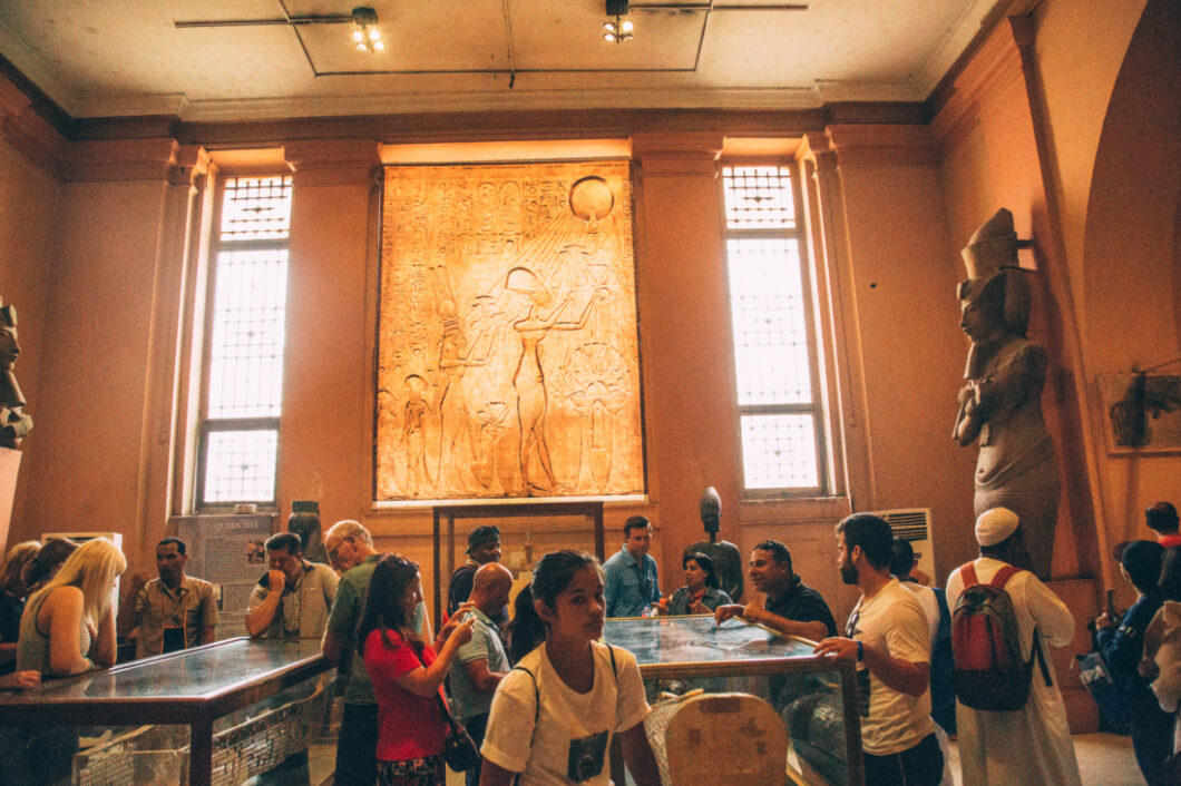 Crowds of museum visitors mill around an exhibit at the Egyptian Museum in Cairo, admiring the artifacts in display cases, statues, and ancient artwork on the walls.