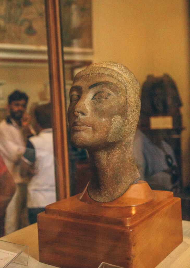 A stone-carved bust status of Nefertiti, the wife of an Egyptian Pharoah, displayed on a wooden stand inside a glass display case.