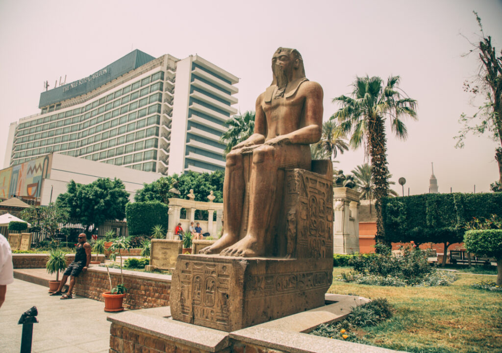 A large stone statue of an Egyptian noble sitting on a throne stands tall in an outdoor space outside of the Egyptian Museum.