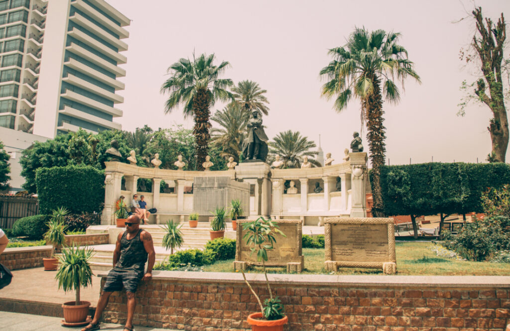 An outdoor space at the Egyptian Museum features trees and manicured landscaping, benches, seating, and ancient structures carved from stone in a courtyard filled with visitors.