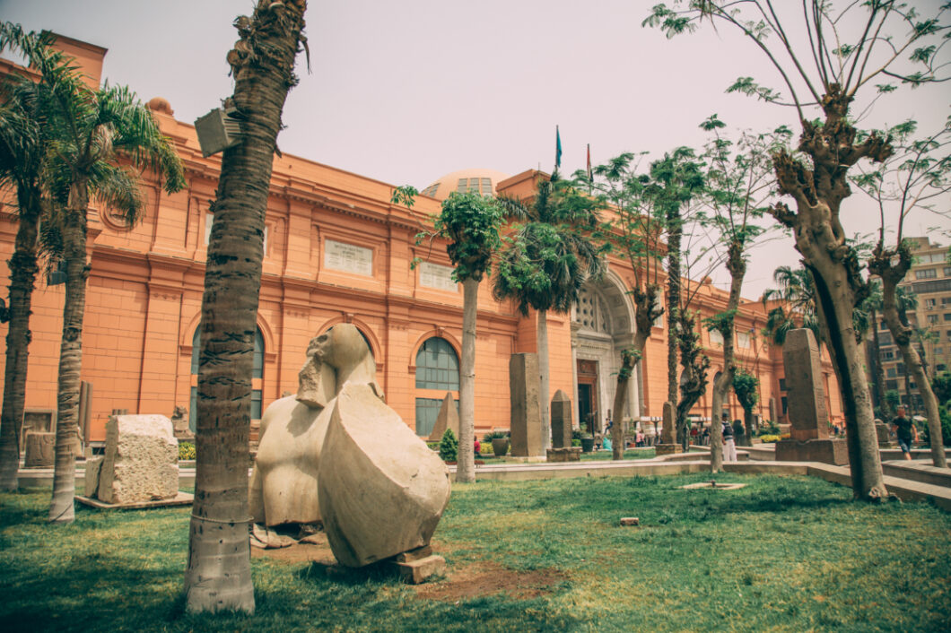 A photo of the outside façade of The Egyptian Museum. The large multi-story building is a terracotta color with windows and arches. A green manicured lawn in front features tall palm trees and sculptures.